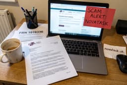 A laptop displaying the FCA website, with a red note saying 'Scam Alert: NovaTask,' papers labeled 'FRN 1016336,' and a coffee cup on a wooden desk.
