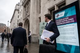 A man in a suit holding documents stands outside the High Court next to a sign about FCA action against Hunter Jones, with people walking by.
